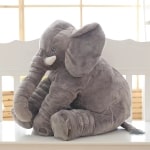 Peluche d'un éléphant gris assis. Il a de grande oreille et des défenses. La peluche en coton est moelleuse.
