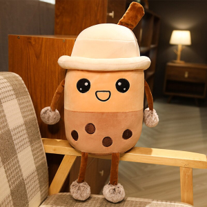 Peluche Bubble tea avec un chapeau , elle est beige et marron, et se trouve assise sur l'accoudoir en bois d'un fauteil à carreaux beige, blanc et marron dans un appartement avec une lampe allumée dans le fond