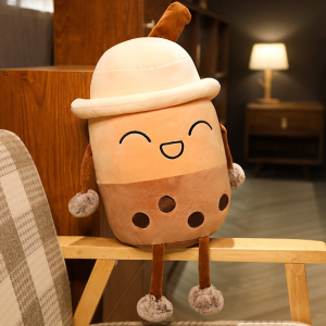 Peluche Bubble tea en train de rire avec un chapeau , elle est beige et marron, et se trouve assise sur l'accoudoir en bois d'un fauteil à carreaux beige, blanc et marron dans un appartement avec une lampe allumée dans le fond