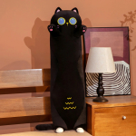 Sur un lit à côté duquel se trouve une table de chevet avec une lampe de chevet blanche et un cadre derrière, se trouve une peluche oreiller de chat noir, il se tient debout sur ses toutes petites pates arrières