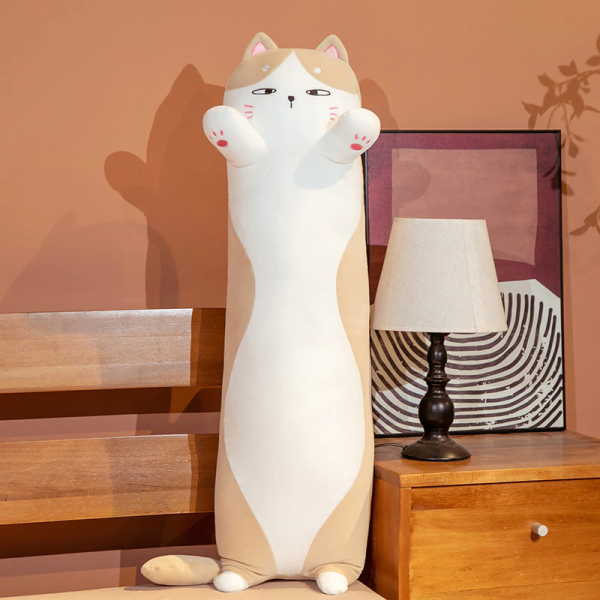 Sur un lit à côté duquel se trouve une table de chevet avec une lampe de chevet blanche et un cadre derrière, se trouve une peluche oreiller de chat blanc et marron, il se tient debout sur ses toutes petites pates arrières