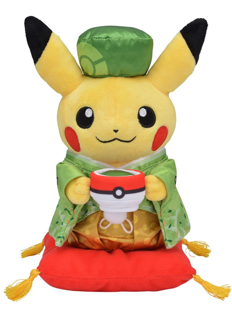 Peluche de pikachu tenant une pokeball et porte une tenue chinoise verte avec un petit chapeau assorti et il se trouve sur un petit coussin rouge