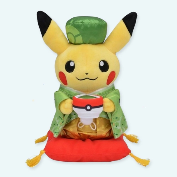 Peluche de pikachu tenant une pokeball et porte une tenue chinoise verte avec un petit chapeau assorti et il se trouve sur un petit coussin rouge