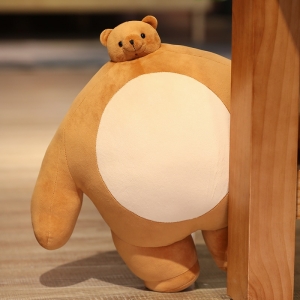 Coussin peluche en forme d'ours de couleur marron avec un ventre beige devant le pied d'un meuble en bois posé sur du parquet en bois