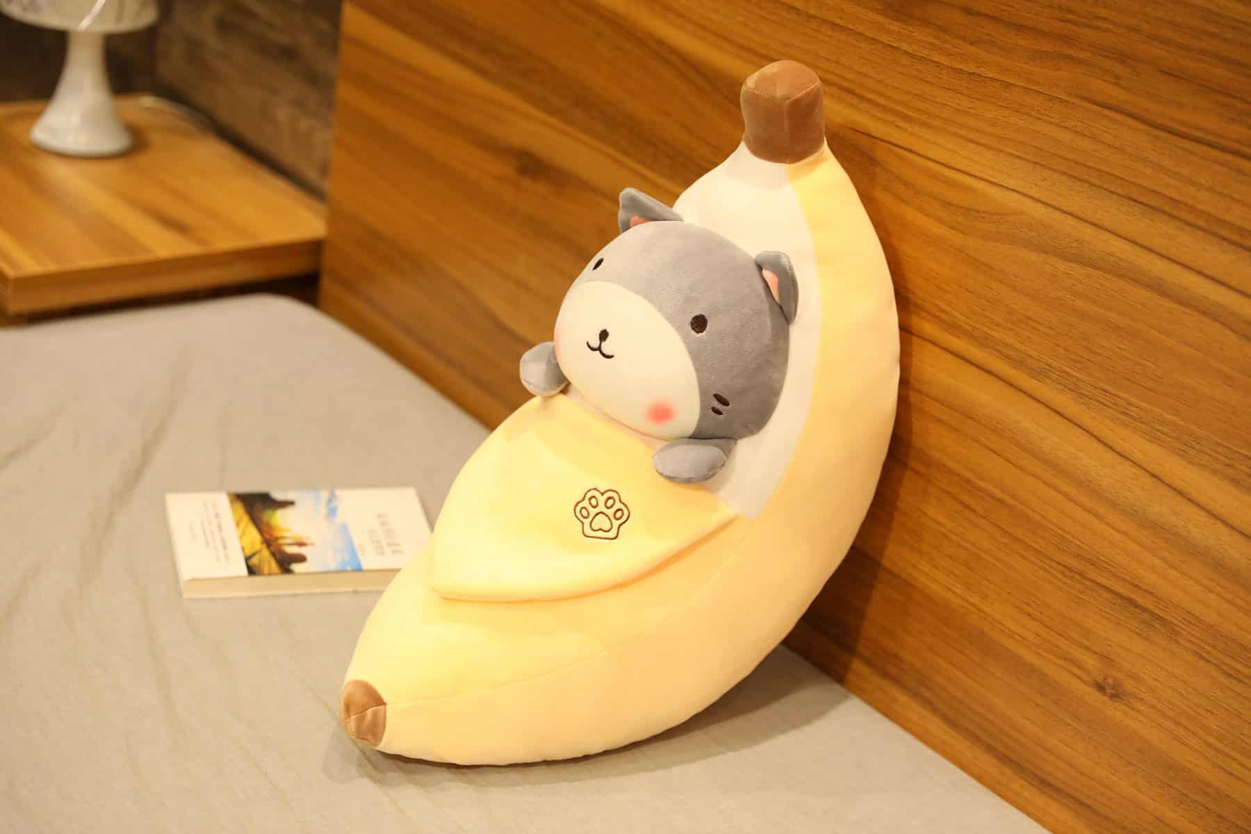 Peluche banane épluchée avec un shiba inu de couleur gris dedans posé sur un lit gris et un contre un mur de couleur bois avec un livre posé à côté de lui